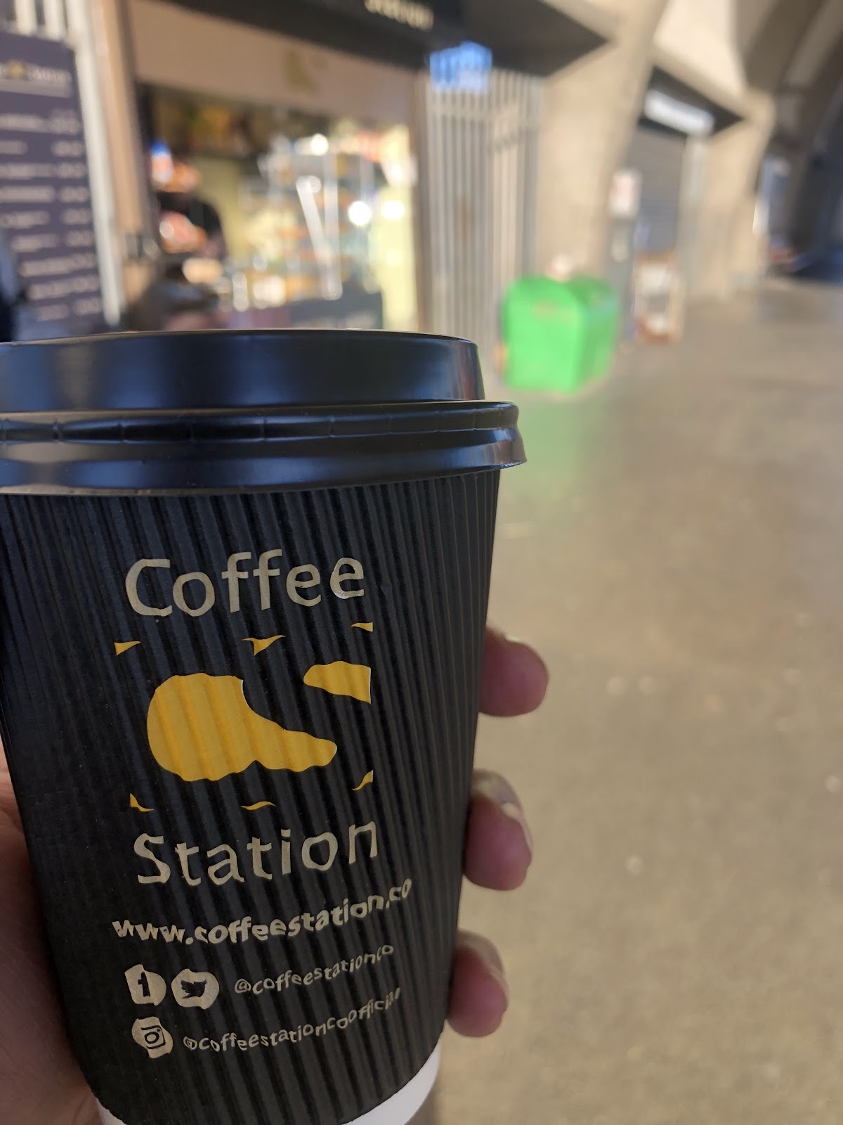 A Coffee Station takeaway cup held up in front of the station concourse
